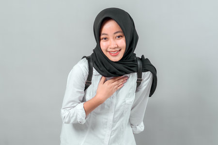 Smiling Muslim teenage girl in hijab with backpack placing hand on chest while slightly bowing. Friendly student greeting gesture for school, education, respect, or gratitude theme.の写真素材