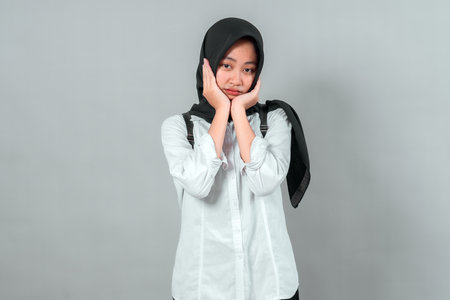Muslim teenage girl in hijab wearing a backpack with hands on cheeks, showing bored or tired facial expression. Back to school concept with student emotion and attitude.の写真素材