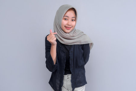 Smiling young Indonesian Muslim woman in a grey hijab and black blouse making a finger heart gesture with one hand, standing confidently against a plain studio background.の写真素材