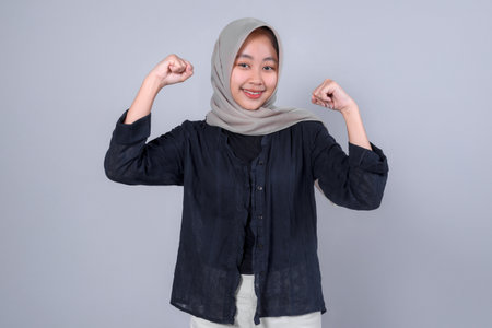 Confident young Indonesian Muslim woman wearing hijab and black shirt smiling while flexing her arms, expressing strength and empowerment on gray background.の写真素材