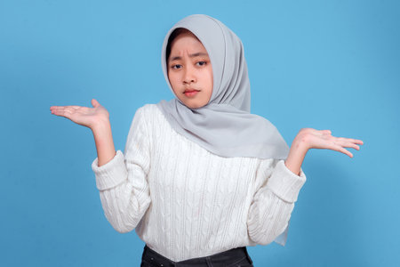Young woman in hijab showing confused or uncertain facial expression, raising both hands in questioning gesture, wearing a white sweater against blue background, looking puzzled.の写真素材