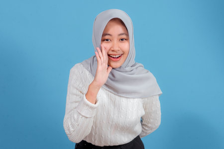 Cheerful young Asian woman wearing a hijab and white sweater smiling happily while cupping her hand to her mouth, as if calling or announcing, isolated on blue background.の写真素材