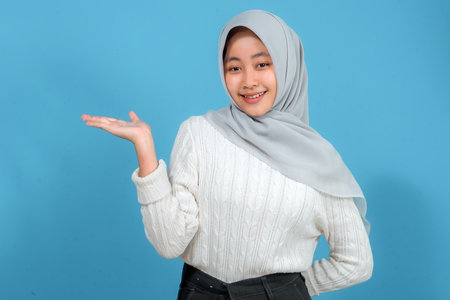 Cheerful young Muslim woman in a grey hijab and white sweater smiling brightly while posing confidently with one hand raised, isolated on a vibrant blue background.の写真素材