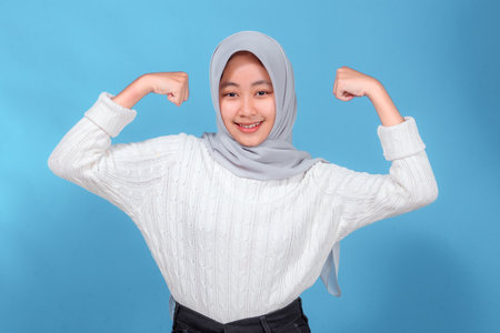 A young woman in a light gray hijab and white sweater smiles confidently while flexing her arms, showing strength, empowerment, and positivity against a bright blue background.の写真素材
