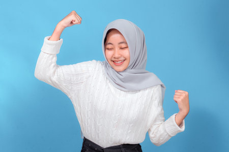 Portrait of a cheerful young woman in a light gray hijab and white sweater, smiling with closed eyes and flexing her arms, showing confidence, strength, and positive energy.の写真素材