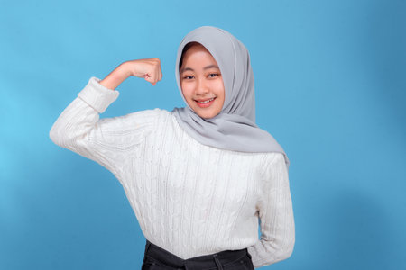 A cheerful young woman wearing a light gray hijab and white sweater confidently poses with a flexed arm, smiling brightly and showing strength against a blue background.の写真素材