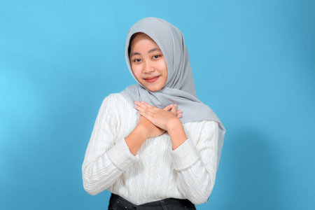 Portrait of a young woman in a light gray hijab and white sweater smiling softly while placing her hands on her chest, expressing gratitude and sincerity with a gentle expression.の写真素材
