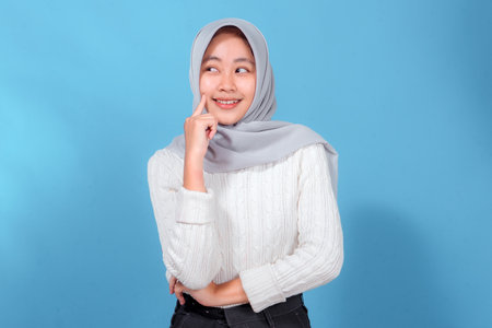 Cheerful young Muslim woman in a grey hijab smiling while touching her cheek with one finger, looking to the side thoughtfully, posing against a bright blue background.の写真素材