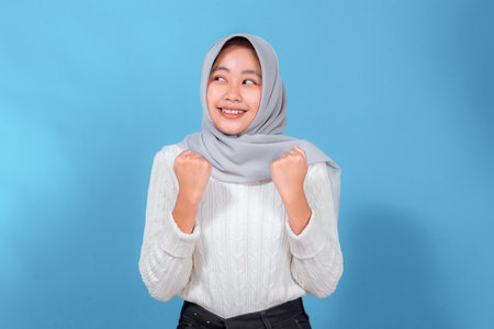 Portrait of a happy young woman in a light gray hijab and white sweater, smiling brightly with clenched fists, showing excitement, enthusiasm, hope, and positive emotions.の写真素材