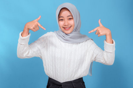 Cheerful young Muslim woman in a hijab smiling brightly while pointing enthusiastically at herself with both hands, standing confidently against a solid blue background. Concept of pride.の写真素材