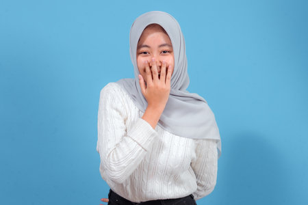 A young Asian woman wearing a light gray hijab and white sweater smiles shyly while covering her mouth with her hand, expressing joy and surprise against a blue background.の写真素材