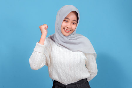 A cheerful young woman wearing a light gray hijab and white sweater smiles confidently while raising her fist in a gesture of success, against a blue background.の写真素材