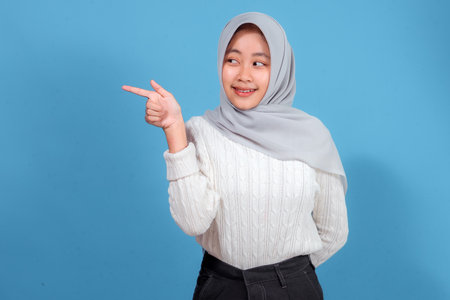 A cheerful young woman wearing a light gray hijab and white sweater is smiling and pointing to the side, standing against a bright blue background, showing curiosity.の写真素材