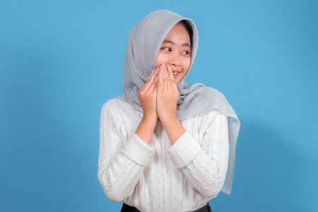 A cheerful young woman wearing a light gray hijab and white sweater smiles shyly while covering her mouth with both hands, expressing happiness and excitement.の写真素材