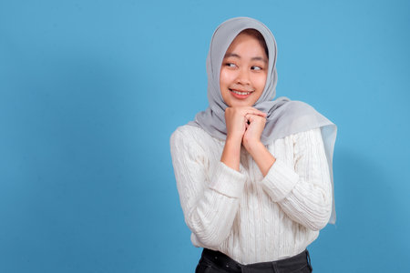 Young Asian Muslim woman with a soft shy smile, wearing a light gray hijab and white sweater, clasping her hands under her chin, isolated on blue background, looking sideways.の写真素材