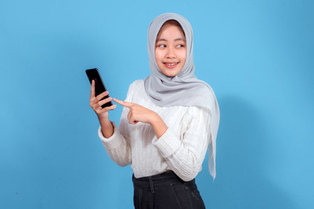 A cheerful young Asian woman wearing a hijab smiling brightly while pointing at her smartphone,...