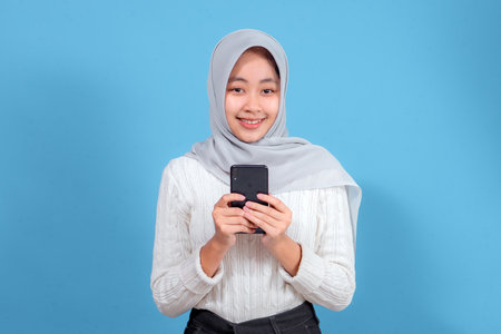 A young woman with a cheerful and happy facial expression, wearing a gray hijab and white sweater,...