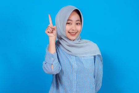 Smiling young woman wearing a hijab and striped shirt confidently pointing upward with one finger,...