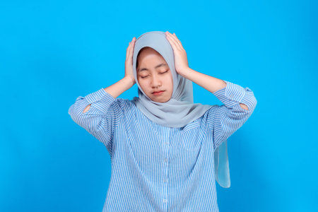 Young Asian woman wearing a hijab and striped shirt showing a stressed or frustrated expression, closing her eyes and holding her head with both hands against a blue background.の写真素材