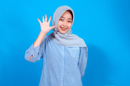 A happy young woman wearing a light gray hijab and striped blue shirt smiling brightly while showing...