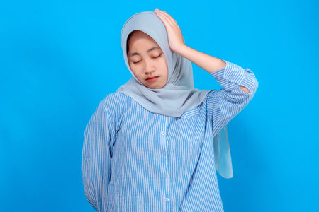 Young Asian woman in hijab and striped shirt expressing sadness and discomfort, closing her eyes and touching her head, standing against a plain bright blue background.の写真素材