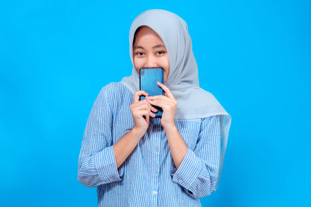 Happy young Asian woman wearing hijab smiling shyly while holding smartphone close to her face,...