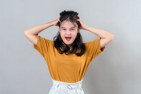 Young Asian woman showing a frustrated and stressed facial expression with hands on head. Her exaggerated face conveys confusion, pressure, or mental overload. Ideal for advertising use.の写真素材