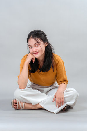 Young Asian woman sitting cross-legged on the floor with a soft, confident smile and relaxed pose. Her expression conveys friendliness, calmness, and thoughtful charm in a studio shot.の写真素材