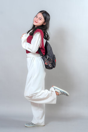 Cheerful young woman smiling brightly with a playful pose, one leg lifted and hands clasped, expressing joy, youthfulness, and genuine happiness in a casual modern look.の写真素材