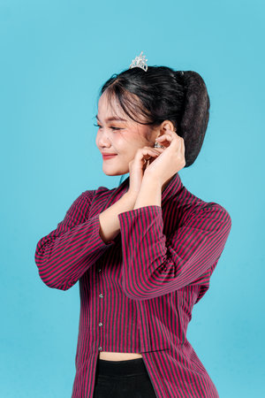 Charming young Asian woman in traditional striped lurik outfit, smiling sweetly while adjusting her earring. Her playful and confident facial expression enhances cultural elegance.の写真素材