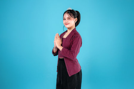 Smiling young Asian woman in traditional striped lurik attire with hands in greeting gesture. Her soft, confident expression radiates warmth, respect, and cultural pride.の写真素材