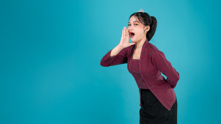 A surprised young woman in traditional Javanese lurik outfit opens her mouth wide with an expressive face, raising one hand beside her cheek against a vibrant turquoise background.の写真素材