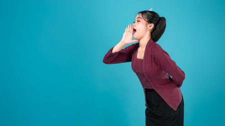 A surprised young woman in traditional Javanese lurik outfit opens her mouth wide with an expressive face, raising one hand beside her cheek against a vibrant turquoise background.の写真素材