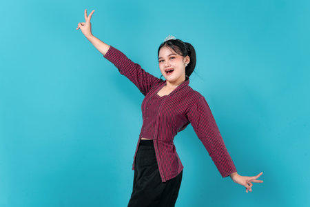 Happy young Asian woman with a tiara showing a wide smile and playful expression while raising both arms in a fun pose. Ideal for lifestyle, joy, and advertisement concepts.の写真素材