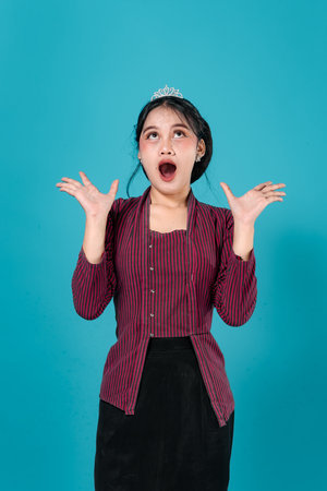 Portrait of a young Asian woman with a tiara showing a dramatic surprised expression, mouth wide open and eyes looking up, standing against a turquoise background in a studio setting.の写真素材