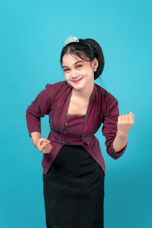 Smiling young Asian woman wearing a traditional outfit and a tiara, confidently making a double fist...