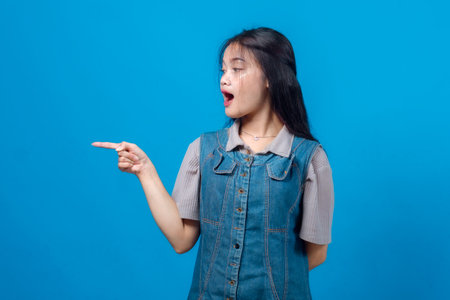 Surprised young Asian woman pointing sideways with an open mouth, wearing a denim dress against a bright blue background. Studio shot for advertising, banner, or promotional design.の写真素材