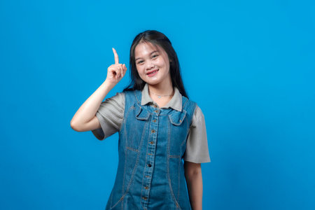 Portrait of a young Asian woman smiling and pointing upwards with her index finger, suggesting an idea or inspiration, against a solid blue background. Cheerful and expressive pose.の写真素材