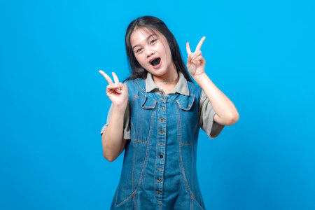 Playful young Asian woman smiling with mouth open and showing double peace signs on both hands. Expressive joyful face, casual denim outfit, vibrant blue background, studio shot.の写真素材