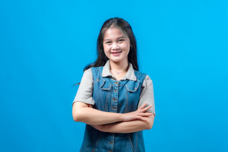 Confident young Asian woman with long black hair smiling and crossing arms, standing against blue...
