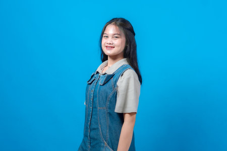 Portrait of a young Asian woman smiling confidently while posing against a solid blue background. She wears a casual denim overall and short-sleeved shirt, looking cheerful.の写真素材