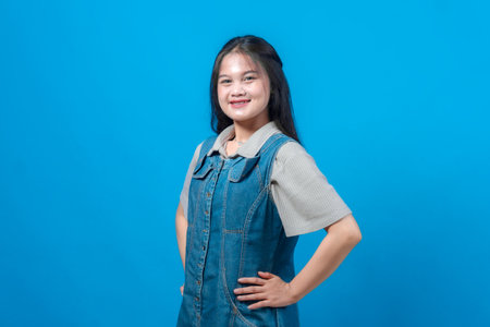Happy young Asian woman smiling confidently with hands on hips, wearing denim dress and standing against solid blue background. Positive expression, casual fashion, studio portrait.の写真素材