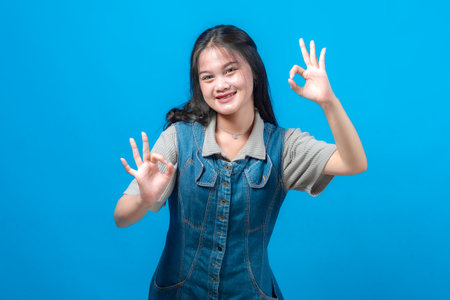 Smiling young Asian woman making double OK hand gestures with confidence, wearing a denim dress and gray shirt, standing against a vibrant blue background in studio lighting.の写真素材