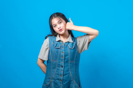 Young Asian woman in a denim dress showing a thumbs up gesture with a playful expression, standing...