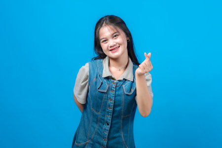 Happy young Asian woman making finger heart gesture with cheerful smile, wearing casual denim dress and standing against bright blue background, symbolizing love and positivity.の写真素材