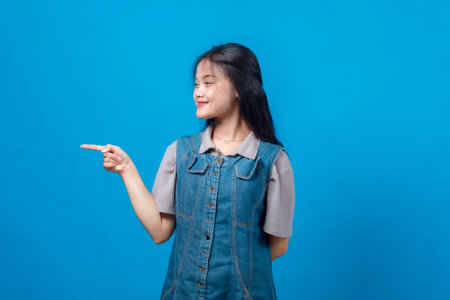 Smiling young Asian woman in a denim dress pointing sideways with her index finger, looking cheerful and confident against a plain blue studio background.の写真素材