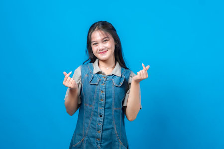 Young Asian woman smiling while making Korean finger heart gesture with both hands, wearing denim dress and gray shirt, isolated on blue background, cheerful studio portrait.の写真素材