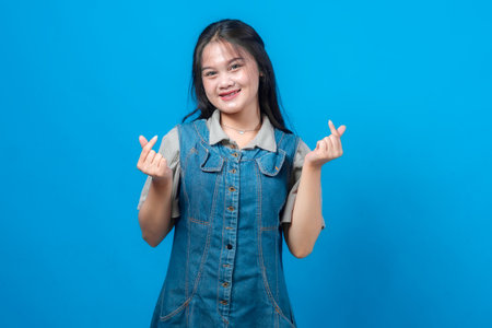 Smiling young Asian woman showing double finger heart gesture with both hands, standing confidently in casual denim dress, isolated on solid bright blue background, studio shot.の写真素材