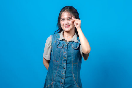 Portrait of happy young Asian woman showing mini heart gesture with fingers near her eye, smiling warmly, wearing denim dress, isolated on blue background, symbol of affection.の写真素材