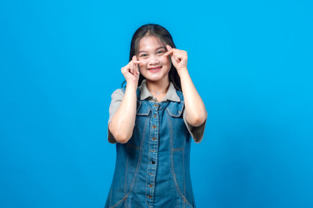 Portrait of cheerful young Asian woman making double finger heart gesture, smiling joyfully, wearing denim dress, isolated on blue background, symbol of love, positivity, and happiness.の写真素材